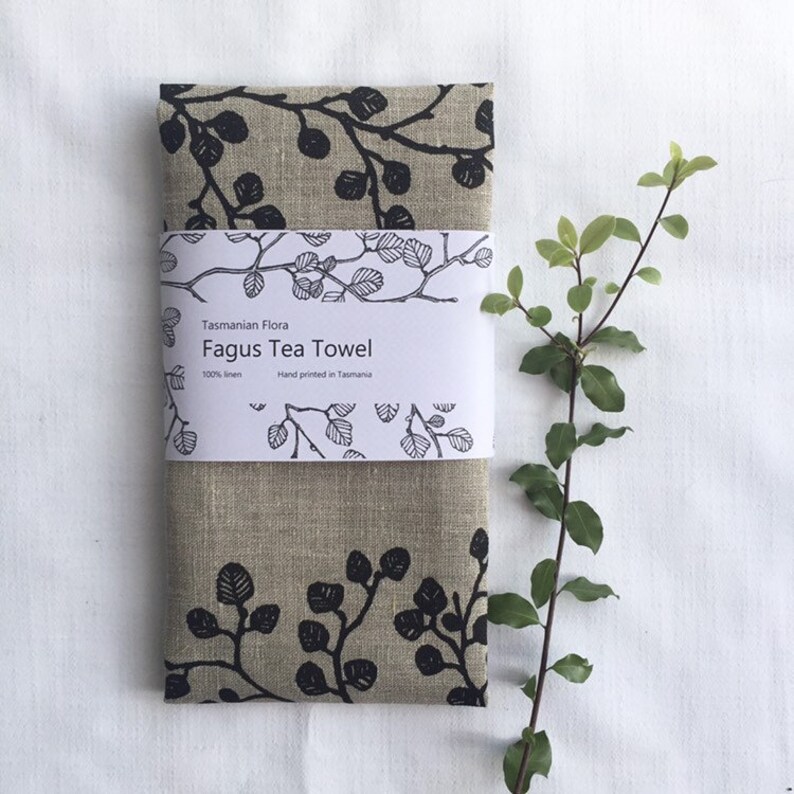 May include: A beige linen tea towel with a black and white print of branches and leaves. The towel has a label that reads "Tasmanian Flora Fagus Tea Towel 100% linen Hand printed in Tasmania."