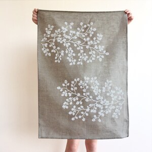 May include: A brown linen tea towel with a white floral design. The design features two branches of leaves and berries.