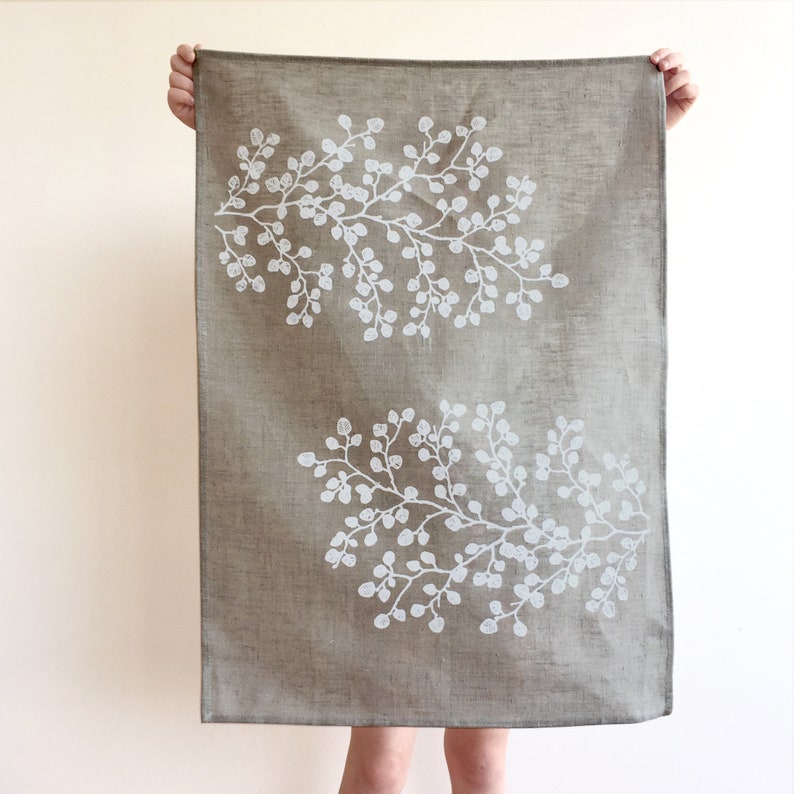 May include: A brown linen tea towel with a white floral design. The design features two branches of leaves and berries.