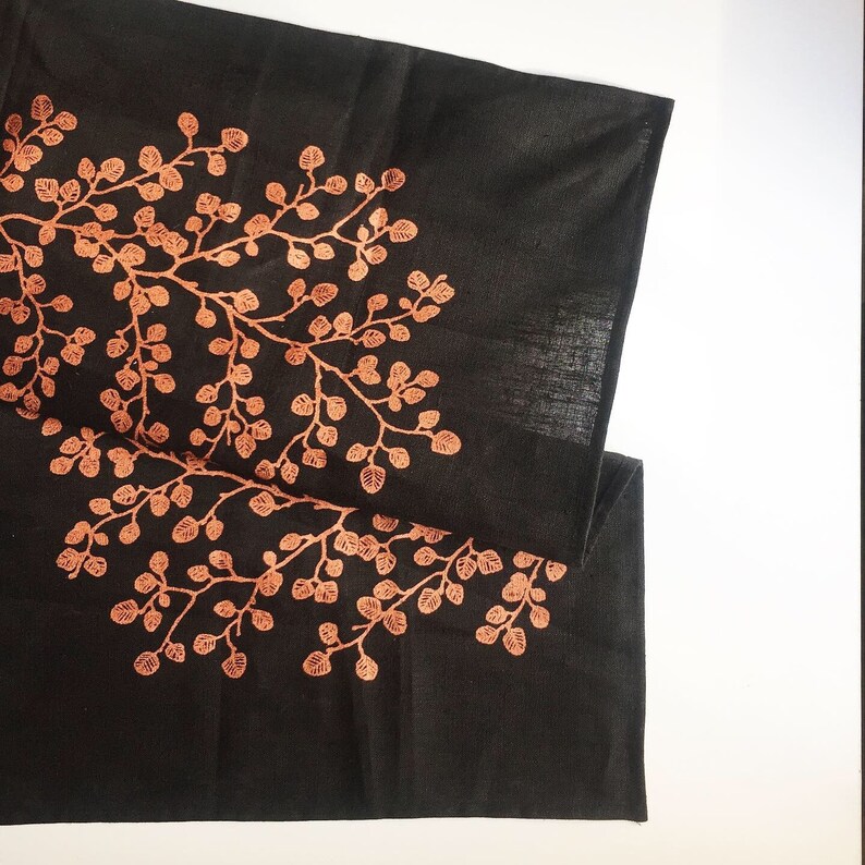 May include: Black linen table runner with a copper-colored floral design. The design features a stylized tree with branches and leaves.
