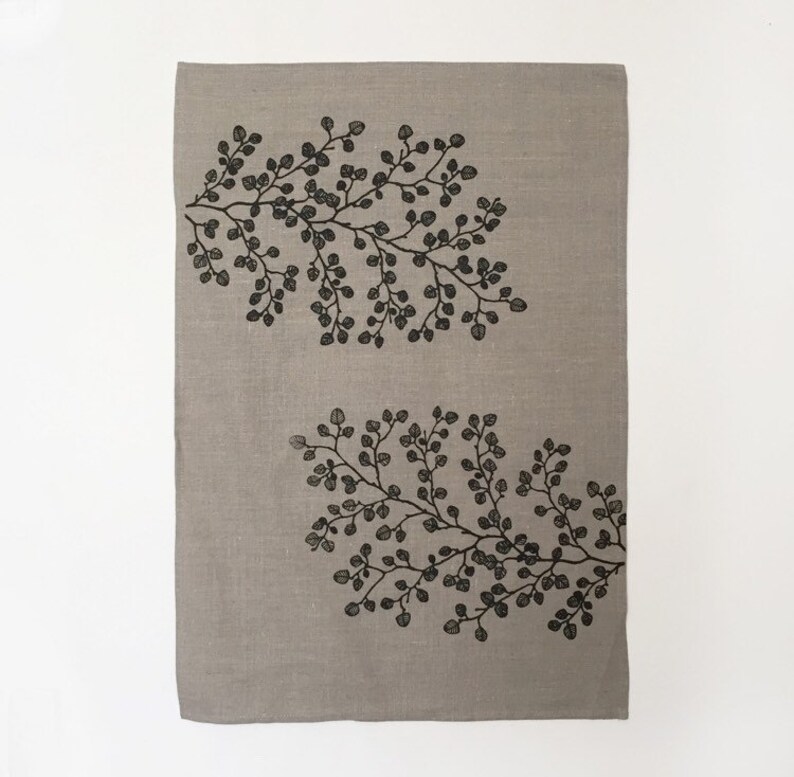 May include: A beige linen tea towel with a black and white floral design. The design features two branches of leaves with small, rounded leaves.