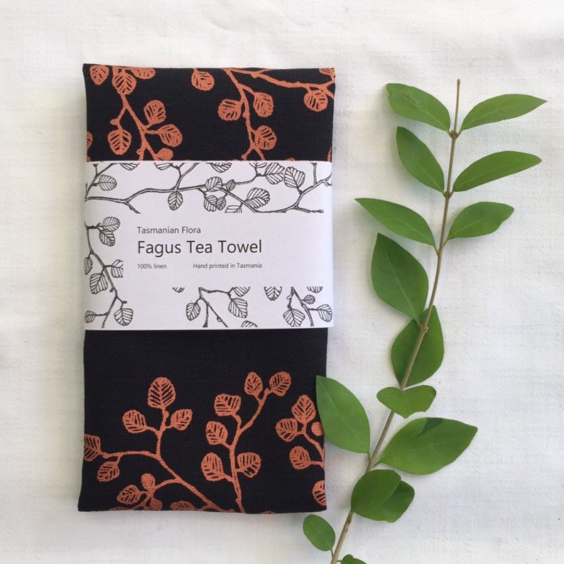 May include: A black tea towel with a copper-colored print of leaves and branches. The towel is labeled "Tasmanian Flora Fagus Tea Towel 100% linen Hand printed in Tasmania."