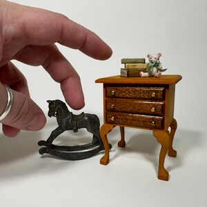 May include: A miniature wooden dresser with three drawers and turned legs, a rocking horse, and a small teddy bear with a green ribbon sitting on top of a stack of books.