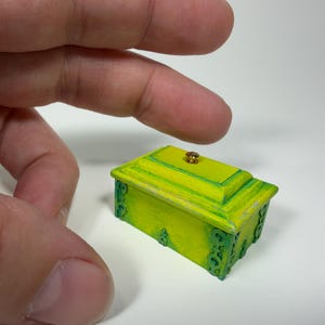 May include: A miniature rectangular box painted in vibrant shades of lime green and yellow. The lid features a small, centered gold-colored knob, and the sides are decorated with intricate, raised green scrollwork patterns along the edges.