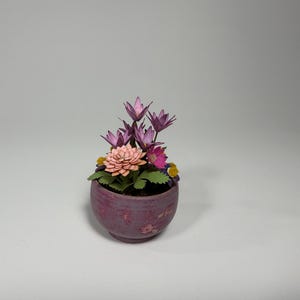May include: A small, round, purple ceramic pot filled with a variety of colorful paper flowers. The arrangement includes pink, purple, and yellow blooms with green leaves. The pot has a textured surface.