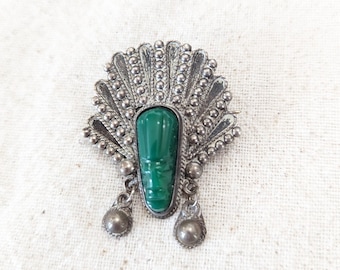 May include: A silver brooch with a green gemstone in the center. The brooch is shaped like a fan with two dangling beads.