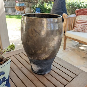 May include: A large, brown ceramic vase with a black base sits on a wooden table. The vase has a smooth, glossy finish and is slightly wider at the top than the bottom.
