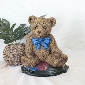 May include: A brown teddy bear figurine with a blue bow tie. The bear is sitting on a base with a green border and a diamond pattern in red, blue, and green. The bear has a textured fur appearance.