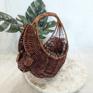 May include: A brown wicker basket with a curved handle. The basket has a unique shape, with a rounded base and a woven design. The basket is sitting on a round, marble-like surface. The basket is a decorative item.