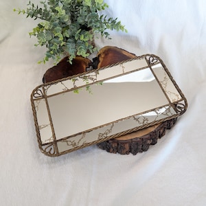 May include: A rectangular vanity tray featuring a reflective mirrored surface with a decorative gold-toned metal frame. The frame has a twisted rope texture and small heart-shaped accents at each corner. The mirror surface displays a subtle marbled pattern.