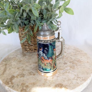 May include: A decorative beer stein with a detailed forest scene featuring deer, trees, and a small cabin. The stein has a silver-colored metal lid and handle. The base is tan, and the body is primarily blue with colorful imagery.