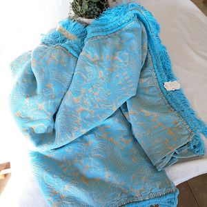 May include: A folded turquoise blanket with a detailed, embossed pattern in a lighter tan color. The blanket has a fringe trim in turquoise and gold accents. The blanket is draped over a white surface.