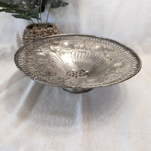 May include: A silver bowl with an intricate embossed design and a small hole in the center. The bowl is on a white surface.