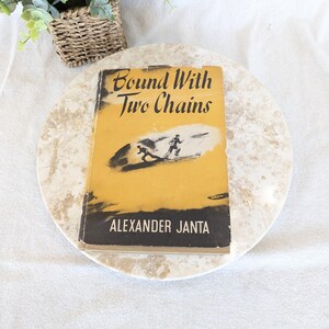 May include: A vintage book titled "Bound With Two Chains" by Alexander Janta. The book has a yellow cover with a black illustration of two figures. The book is placed on a marble-like surface.