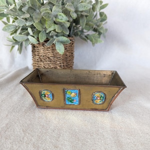May include: A small, rectangular brass planter with a copper rim and three blue and yellow enamel floral designs.