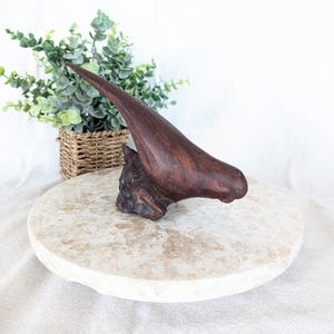 May include: A dark brown, abstract wooden sculpture with a pointed, curved shape. The sculpture is mounted on a light-colored, round stone base. A woven basket with green foliage is in the background.