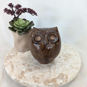 May include: A brown ceramic owl figurine with large, round eyes. The owl is sitting on a beige marble-like surface.