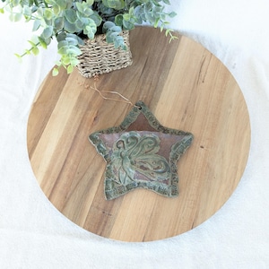 May include: A star-shaped ceramic ornament with a green and brown design. The ornament features a central image of a stylized figure and decorative border. It is displayed on a wooden cutting board with a small woven basket and greenery.