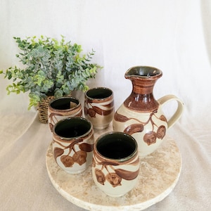 May include: A ceramic pitcher and four matching cups with a brown cherry design. The pitcher has a handle and a spout. The cups are cylindrical with a dark green interior. The set is displayed on a round, marble-like tray.