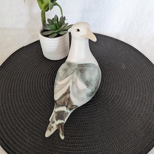 May include: A ceramic dove figurine with a white body and gray and brown wing details. The bird has a beige beak and is positioned on a black circular mat. A small potted plant with green leaves is in the background.