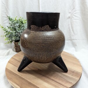 May include: A brown, three-legged, antique-style vase with etched designs. The vase has a dark brown rim and sits on a wooden round tray. A small green plant in a basket is in the background.