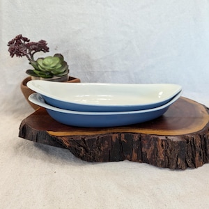May include: Two oval-shaped serving dishes. The dishes are blue on the outside and white on the inside. They are stacked on top of each other. The dishes are on a dark brown wooden surface.