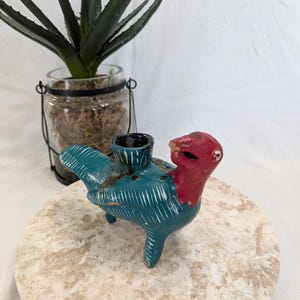 May include: A small, hand-painted ceramic bird figurine featuring a vibrant teal body with white etched line details and a solid red head. The bird has a cylindrical opening on its back and stands on three short legs, showing signs of wear.