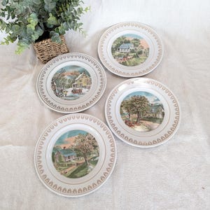 May include: Four vintage decorative plates with scenic landscape illustrations. Each plate features a different rural scene with houses, trees, and seasonal elements. The plates have a gold trim and a cream-colored background.