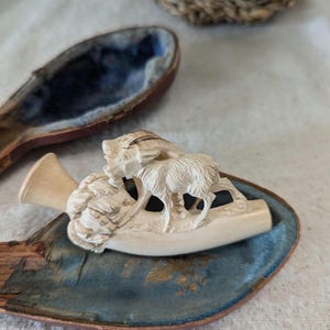 May include: A white carved pipe with a depiction of two goats in a mountainous landscape. The pipe is in a blue velvet lined case.