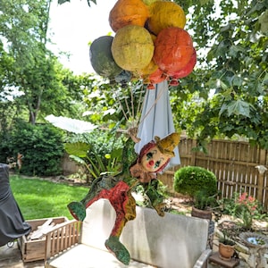 May include: A whimsical clown doll hangs from a cluster of colorful balloons. The balloons are painted in shades of green, orange, and yellow. The clown is dressed in a green, red, and yellow outfit with a painted face and a yellow hat.