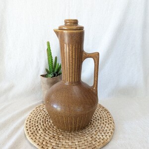 May include: A brown ceramic pitcher with a lid and a handle. The pitcher has a textured surface and is sitting on a woven mat.