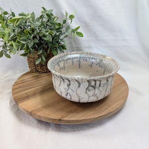 May include: A ceramic bowl with a crackle glaze pattern in shades of white and gray. The bowl sits on a round wooden serving board. A small woven basket with green foliage is to the left of the bowl.