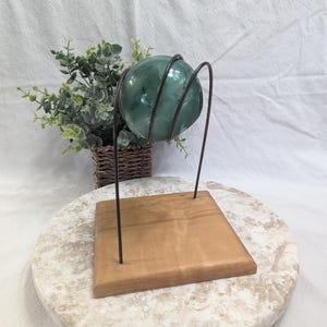 May include: A translucent green glass sphere rests within a dark metal wire frame stand. The stand is attached to a square, light-colored wooden base. The sphere is positioned above the wooden base, held securely by the curved metal wires.