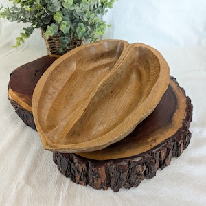 May include: A wooden, heart-shaped serving bowl with a central divider, resting on a dark, round wooden slab with a natural bark edge. The bowl is light brown, and the wood slab is dark brown. A small green plant in a woven basket is in the background.