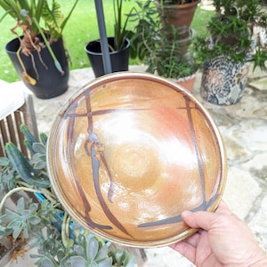 May include: A round ceramic bowl with a glossy, warm-toned glaze in shades of tan, amber, and soft orange. The interior surface features dark brown, hand-painted linear brushstrokes that intersect to form a geometric pattern across the base.