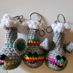 May include: Three handmade crochet keychains shaped like bongs. Each gray bong has a rainbow stripe, black button eyes, and a smiling mouth. The top of each bong is white and fluffy, and each has a silver keyring.
