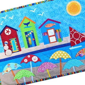 May include: A colorful quilted wall hanging featuring a beach scene with beach houses, a sailboat, a snowman, and umbrellas. The background is blue with white polka dots.