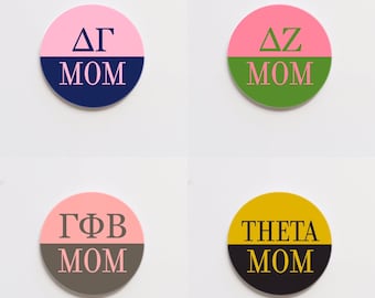 Può includere: Quattro adesivi rotondi bicolore con lettere greche e la parola "MOM" stampata su di essi. Gli adesivi sono rosa e blu scuro, rosa e verde, rosa e grigio e giallo e nero.
