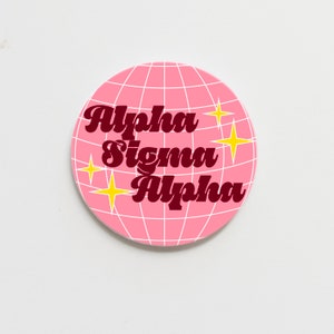 May include: Pink and white round coaster with a grid pattern and the text "Alpha Sigma Alpha" in maroon with yellow stars.