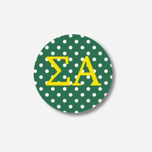 May include: A circular sticker with a dark green base and white polka dots. The sticker displays the Greek letters Sigma and Alpha in a bright yellow colour.