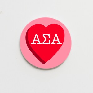 May include: A circular, pink sticker featuring a red heart at its centre. The heart displays the white Greek letters AΣA. The sticker has a light pink border and is set against a white backdrop.