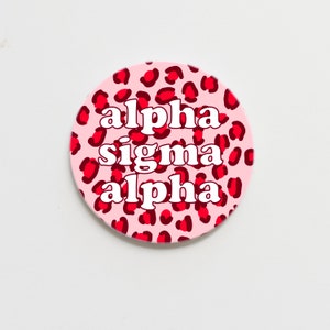 May include: A pink and red round sticker with a leopard print pattern. The text "alpha sigma alpha" is written in white letters on the sticker.