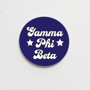 May include: A blue round button with white text that reads "Gamma Phi Beta" with two white stars on either side of "Phi".