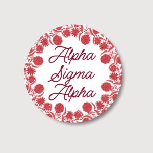 May include: A circular sticker featuring the text "Alpha Sigma Alpha" in a burgundy script font. The text is set against a white backdrop and framed by a red floral wreath.
