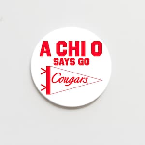 May include: A white round button with red text that says "A Chi O Says Go Cougars" with a red pennant shape below the text.