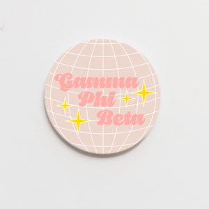 May include: A pink and white round coaster with a grid pattern and the text "Gamma Phi Beta" in pink with yellow stars.
