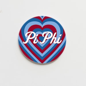 May include: A round button with a blue and red heart pattern. The text "Pi Phi" is written in white cursive letters on the button.