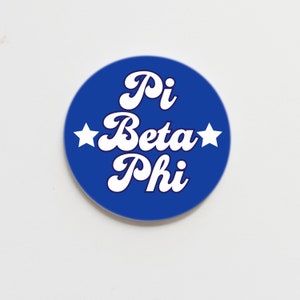 May include: A round, blue button with the words "Pi Beta Phi" in white, stylized script. Two white stars flank the words. The button is set against a white background.