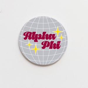 May include: A round, grey, disco ball-style coaster with a white grid pattern. The words "Alpha Phi" are written in a burgundy, retro font. There are four yellow stars on the coaster.