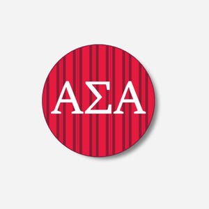 May include: A circular red badge featuring vertical stripes and the Greek letters ΑΣΑ in white. The badge has a white border and a subtle shadow, creating a 3D appearance. The design is clean and striking.